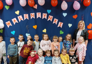 Zdjęcie przedstawia grupę dzieci wraz z wychowawcą. Wszyscy stoją na tle dekoracji walentynkowej.